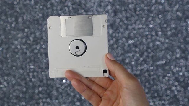 Female hand holds up white floppy disk. Obsolete computer technology. Silver background. Note: Writing on disk says 'Made in Canada'. 1980s. 1990s.