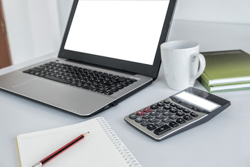 Laptop white screen ,calculator white screen ,white coffee mug, green book and a pencil placed over notebook. All on the white desk