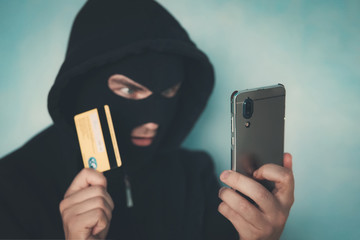 Close up of a man in robbery mask and hood holding the credit card and looking at the smartphone...