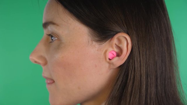 Attractive caucasian woman inserts pink plug into ear for hearing protection. Closeup. Green screen background.