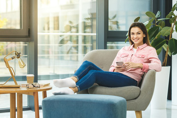 Searching more information. Beautiful young smiling woman looking at her smart phone while sitting in a big comfortable chair at home.