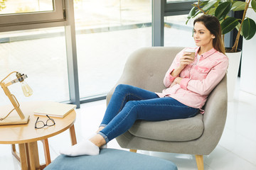 Young smiling woman sitting on sofa while drinking hot coffee. Young brunette woman thinking at home in a leisure time. Happy girl relaxing at home on big chair.