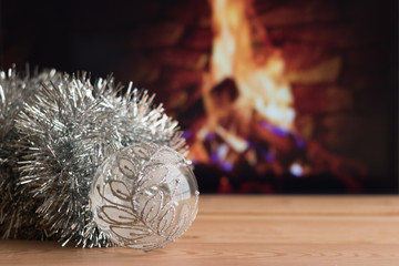 Close-up: Christmas decorations of silver glassy ball and tinsel are on a wooden table on a blurred background of burning fire in fireplace. Concept: Merry Christmas and Happy New Year holidays.