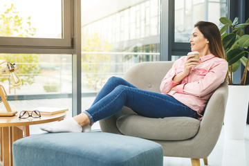 Young smiling woman sitting on sofa while drinking hot coffee. Young brunette woman thinking at home in a leisure time. Happy girl relaxing at home on big chair.