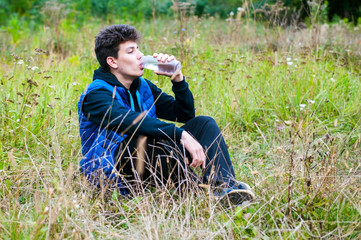 Naklejka na ściany i meble A beautiful teenager sitting on the grass and drinking water from the bottle after training in the forest in the fresh air