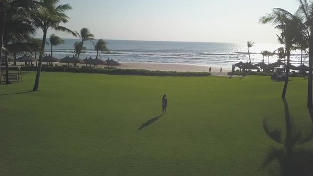 2K Aerial view of a girl walking barefoot across the green grass lawn near the lonely beach seashore and the ocean among the palm trees in luxury resort at sunrise. 