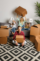 Fototapeta premium Photo of man sitting on floor among cardboard boxes and woman with son and daughter sitting on gray sofa