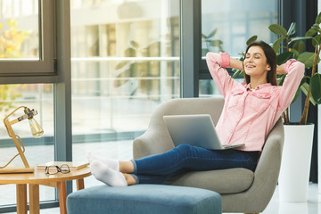 Obraz premium Enjoying relaxing time after work at home. Beautiful young smiling woman working on laptop and drinking coffee while sitting in a big comfortable chair at home. Freelance concept.