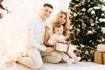 Portrait of beautiful young family on Christmas tree and white cotton background. Attractive parents and a little son open new year gifts