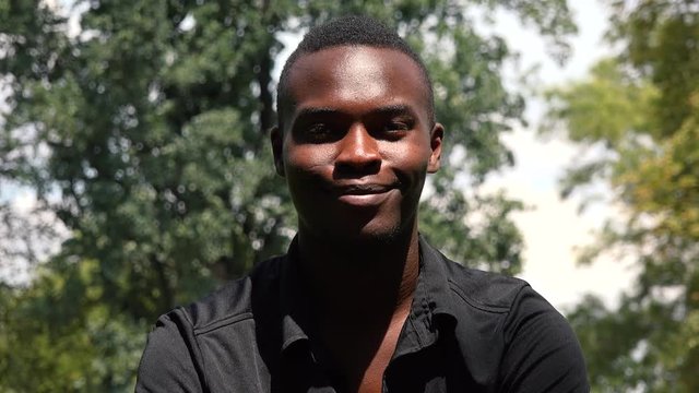 A young black man sits on grass in a park and smiles at the camera - closeup
