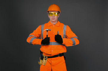 Confident builder worker in a hardhat is holding in hands a hammer and pliers isolated on gray background. Under construction concept.