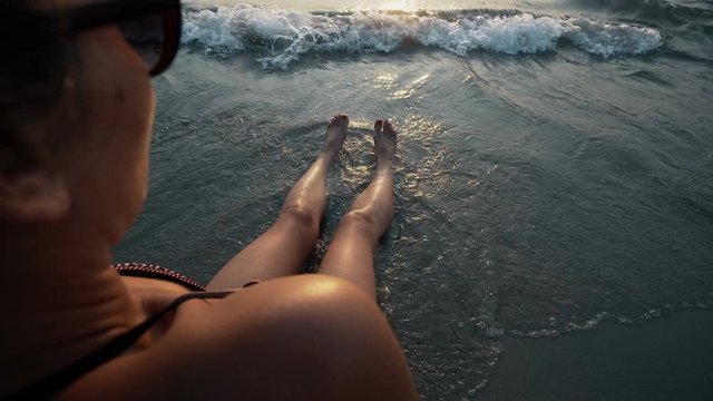 Woman legs and bare feet sitting on golden sand beach at sunset and looking at ocean water seaside