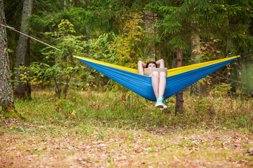 Image of woman with hands behind head resting in hammock with laptop in forest