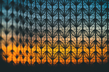 Wonderful vivid dawn from window through patterned curtain. Amazing warm sky behind silhouettes of tulle texture. Orange sunlight. Cosiness textured colorful background with sunrise. Copy space.