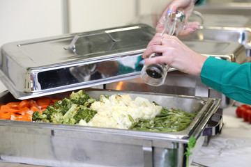 Kelnerka posypuje pieprzem gotowane warzywa na szwedzkim stole w czasie kateringu.	