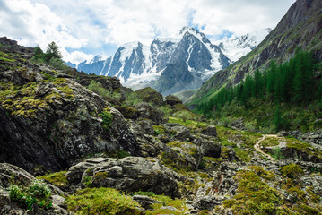 Snowy mountain top behind rocky mountains under cloudy sky. Rocky slope above forest. Clouds above glacier. Atmospheric landscape of majestic nature of highlands. Footpath through stones.