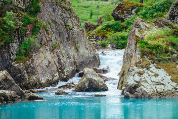 Mountain creek flows into azure lake. Fast stream of brook. Smooth water surface. Wonderful rocks with rich vegetation of highlands. Amazing landscape of majestic nature.