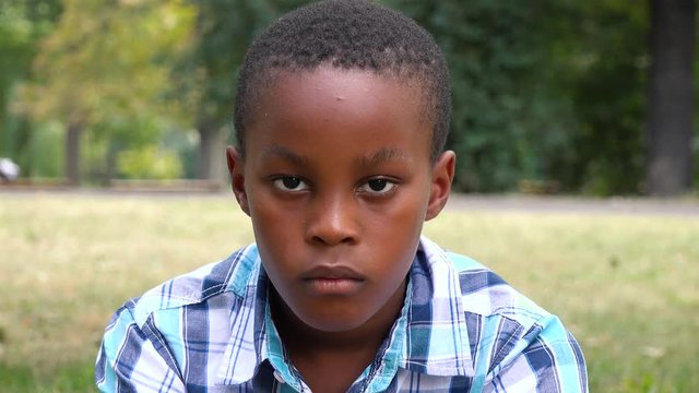 A young  black boy sits on grass in a park and looks seriously at the camera - face closeup