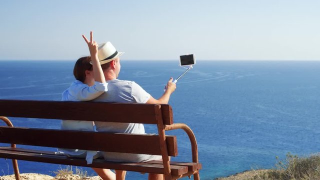Young couple in love relaxing on bench at edge of cliff doing selfie on mobile phone with selfie stick. Husband and wife on honeymoon enjoying view of blue Mediterranean sea making photos on cellphone