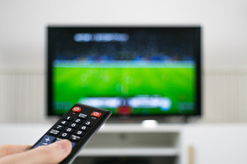 Hand holding TV remote control in front of blurred TV background