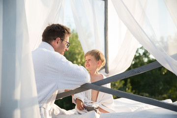 The cute happy little boy sitting on a bed outdoor and looking at his father who is standing near