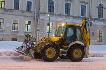 Yellow tractor clears the road and a sidewalk after snowfall. City building at background.