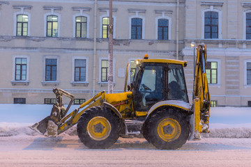 Obraz na płótnie Canvas Yellow tractor clears the road and a sidewalk after snowfall. City building at background.