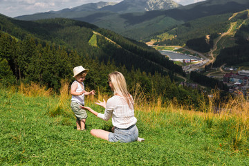 Happy cute little boy standing near his mother who is sitting on the grass against the background of the mountains