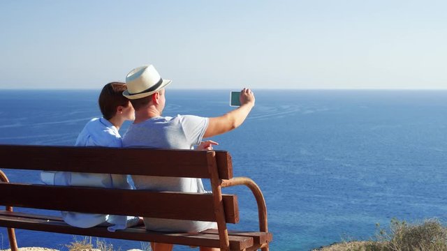 Young couple in love relaxing on bench at edge of cliff taking pictures on mobile phone. Husband and wife on honeymoon enjoying breathtaking view of blue Mediterranean sea making photos on cellphone