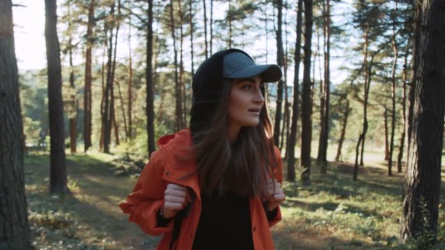 Young woman in cap walking in forest on sunny autumn day look around smile adventure nature outdoor sunlight enjoying freedom sky sunshine vacation wind backpack trip travel slow motion close up