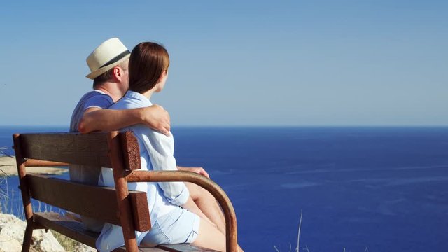 Young couple in love sitting relaxing on bench at edge of cliff. Husband in hat and wife on honeymoon hugging enjoying breathtaking view of blue Mediterranean sea. Concept of freedom and love