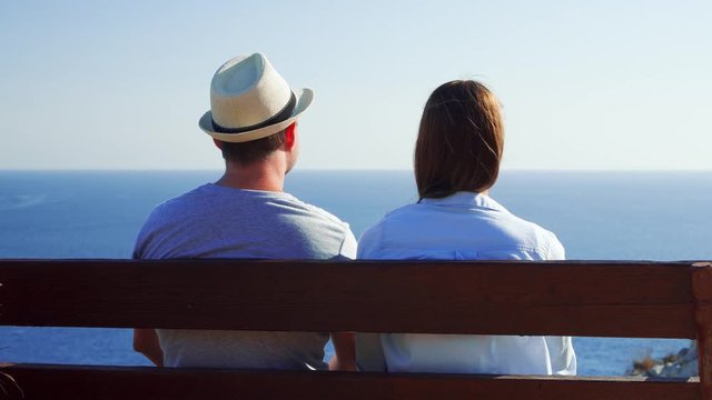 Young couple in love sitting relaxing on bench at edge of cliff. Husband in hat and wife on honeymoon enjoying breathtaking view of blue Mediterranean sea. Concept of freedom and love