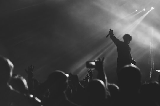 Black and white photo of frontman silhouette in a stage backlights with hand with microphone raised up to the crowd of people