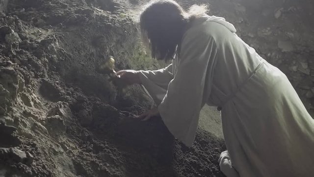 A man like Jesus Christ is digging a hole in the dungeon, a man is searching for a precious treasure, a man in a white robe is digging the earth with a stick and finds gold, Jesus hides the holy grail