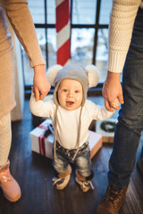 Fototapeta na wymiar Funny little baby boy 1 year old learning walk home in winter in a decorated New Year house. Young family dad and mom hold by the hands of his son in the loft interior wooden floor near the window
