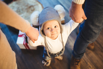 Funny little baby boy 1 year old learning walk home in winter in a decorated New Year house. Young family dad and mom hold by the hands of his son in the loft interior wooden floor near the window