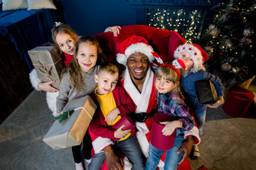 Beautiful children with gifts in their hands around African Santa sitting in an armchair on the background of a Christmas tree