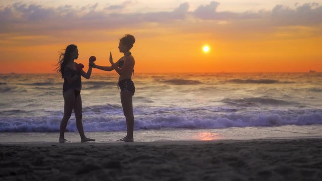 Two girls exercise, training karate on ocean beach against sunset sky and rolling waves