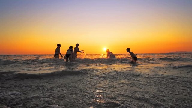 Group of happy people playing and spraying in water at the beach on sunset. Beauty and joyful teenager friends having fun, dancing, spraying over summer sunset. Beach party. Sun flare. Slow motion