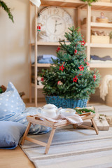 Christmas decoration in living room, tea time. Wooden tray under Christmas tree with red ornaments. Serving morning breakfast with white teapot, mug of tea, blue pillow on floor in cozy home interior