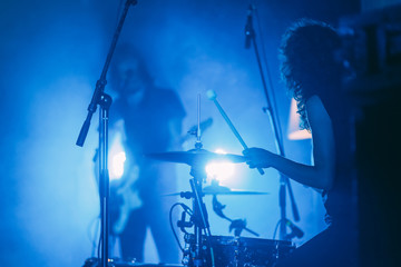 Female drummer with long curly hair in front of singing guitarist on a stage in a bright blue lights playing rock show