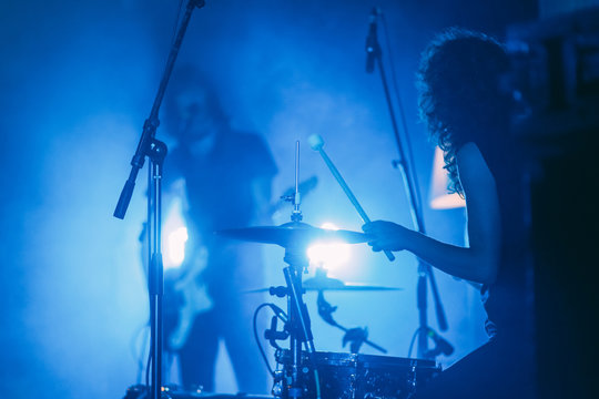  Female drummer with long curly hair in front of singing guitarist on a stage in a bright blue lights playing rock show - Powered by Adobe