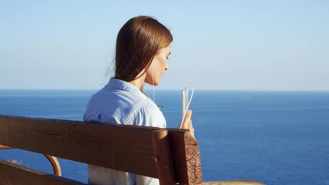 Young woman sitting relaxing on bench at edge of cliff listening music with earphones via app on mobile phone. Female enjoying breathtaking view of blue Mediterranean sea