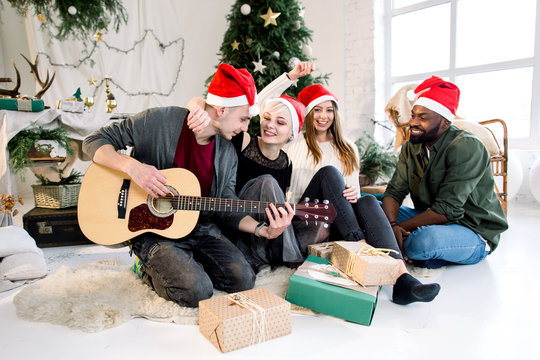Picture showing group of four friends celebrating Christmas at home. Young Caucasian man is playing guitar and the girls and African man are smiling and singing carols
