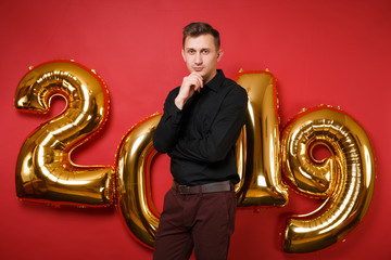 Merry confident young man in black shirt celebrating holiday party standing isolated on bright red wall background, golden numbers air balloons studio portrait. Happy New Year 2019 Christmas concept.