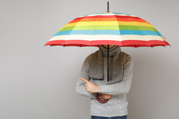 Young man in gray sweater, scarf covering face with colorful umbrella, pointing index finger aside...