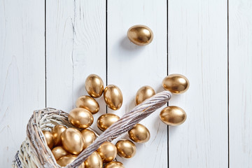 Easter golden eggs spilled out of wicker basket on white wooden table background. Top view