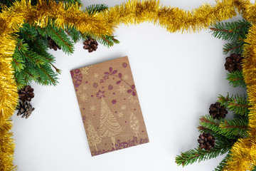 Fototapeta premium Christmas greeting card is lying on white table, surrounded by festive decorations like tree branches, cones and gold tinsel. Merry Christmas.