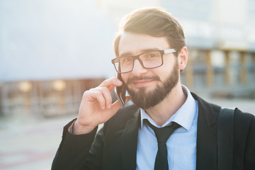 Portrait of smiling bearded businessman in suit using smartphone on office build and sunlight background.