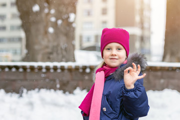 Portrait of happy adorable child showing OK sigh. Cute little caucasian girl in knitted hat and scarf and dawn jacket having fun playing outdoor in winter. Weather and seasons